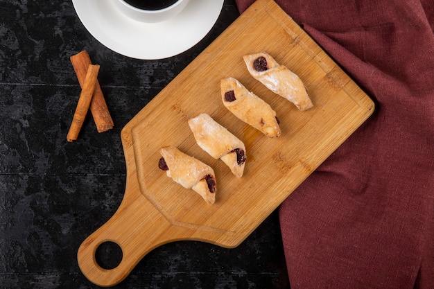 Vista superior de biscoitos de farinha com geléia de morango em uma placa de madeira com uma xícara de café e paus de canela em fundo preto