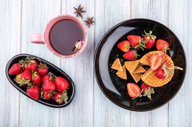 Vista superior de biscoitos waffle com morangos em pratos e xícara de chá na superfície de madeira