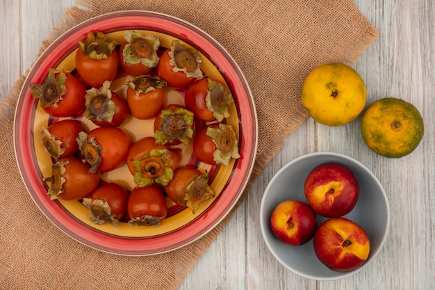 Foto grátis vista superior de caquis frescos em um prato em um pano de saco com pêssegos em uma tigela com tangerinas isoladas em uma parede de madeira cinza