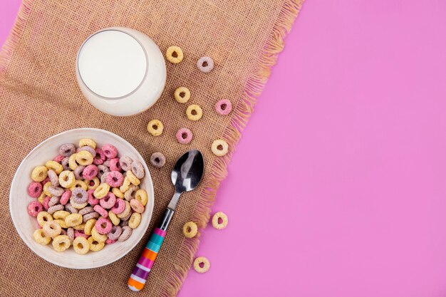 Foto grátis vista superior de cereais multicoloridos em uma tigela com um copo de leite com uma colher com cereais isolados no pano de saco na superfície rosa