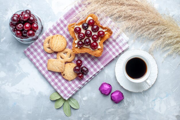 Vista superior de cerejas frescas dentro do prato com bolo cremoso em forma de estrela, chá e biscoitos na mesa branca, biscoito de bolo azedo de frutas