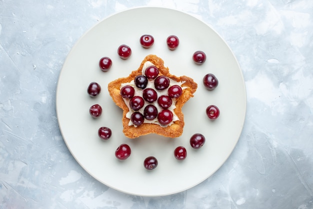 Vista superior de cerejas frescas dentro do prato com bolo em forma de estrela em uma mesa branca clara, vitamina de frutas azedas verão