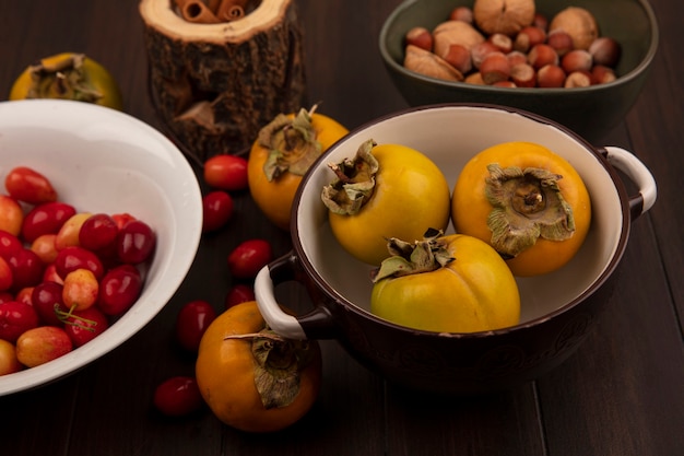 Vista superior de deliciosas cerejas da cornalina em uma tigela branca com frutas de caqui em uma tigela com nozes em uma tigela com paus de canela em uma jarra de madeira sobre uma superfície de madeira