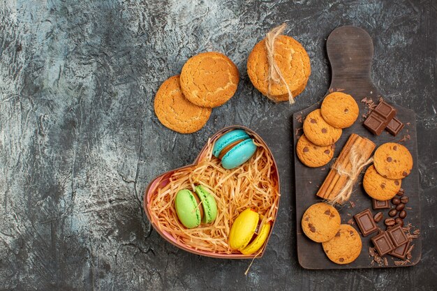 Vista superior de deliciosos biscoitos, barras de chocolate e macarons em uma caixa em forma de coração no fundo escuro glacial