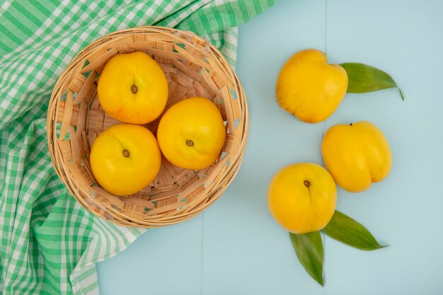 Vista superior de deliciosos pêssegos amarelos frescos em um balde em uma toalha de mesa quadriculada em um fundo azul