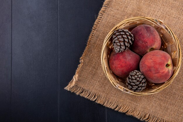 Vista superior de deliciosos pêssegos com pinhas no balde no pano de saco no preto com espaço de cópia