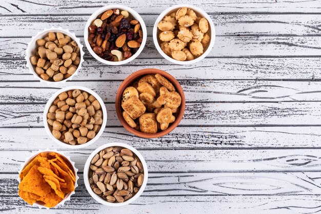 Vista superior de diferentes tipos de lanches como nozes, bolachas e biscoitos em tigelas com espaço de cópia na superfície de madeira branca horizontal