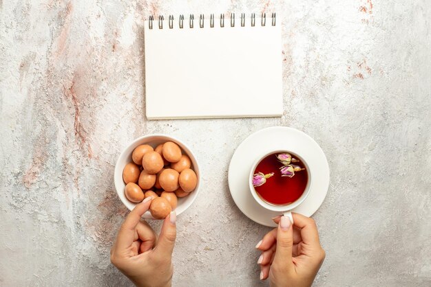 Vista superior de doces à distância com uma xícara de chá branco caderno ao lado da tigela de doces e uma xícara de chá na mão na superfície branca