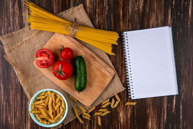 Vista superior de espaguete cru com tomate e pepino em uma tábua com um caderno em um guardanapo bege em uma superfície de madeira