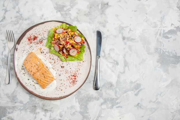 Vista superior de farinha de peixe e salada deliciosa em um prato e talheres colocados no lado esquerdo em uma superfície branca manchada