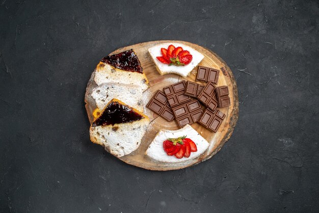 Vista superior de fatias de bolo saborosas com morangos e barras de chocolate na superfície escura
