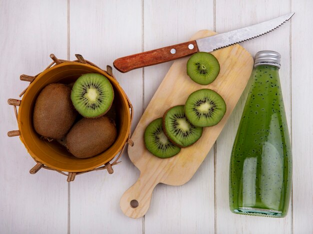 Vista superior de fatias de kiwi com faca na tábua com garrafa de suco na parede branca