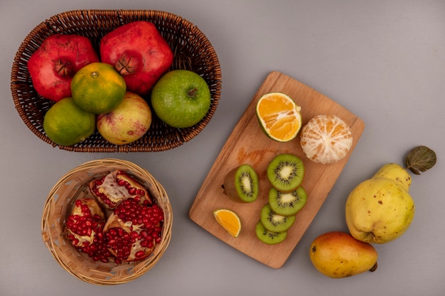 Foto grátis vista superior de fatias de kiwi fresco picado em uma placa de cozinha de madeira com tangerinas e romãs em um balde com pêra e marmelo isolado