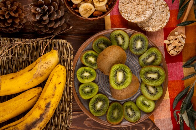 Vista superior de fatias de kiwis em um prato e cacho de bananas em uma cesta de vime, colher de pau com amendoim e bolachas de arroz na madeira