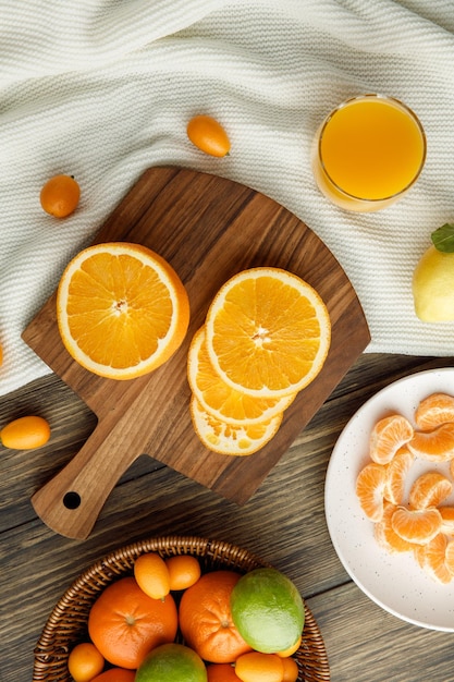 Vista superior de fatias de laranja na tábua com tangerinas kumquats limões na cesta com fatias de tangerina no prato e suco de laranja em pano em fundo de madeira