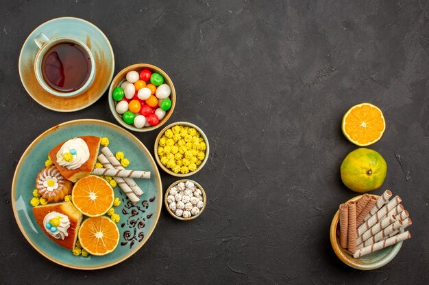 Vista superior de fatias de torta deliciosas com tangerinas frescas e uma xícara de chá na mesa escura