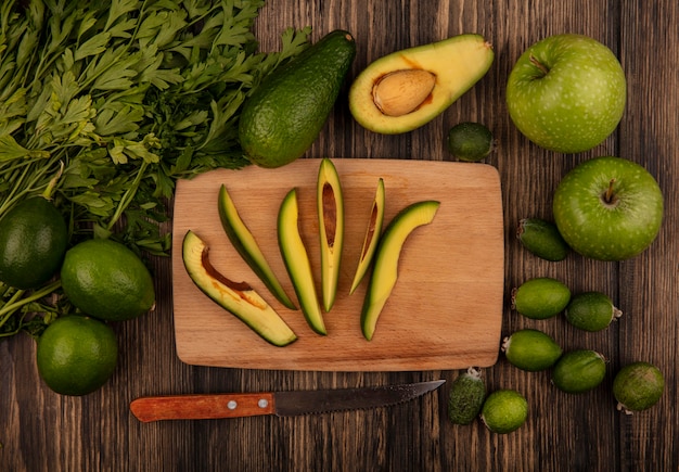 Vista superior de fatias frescas de abacate em uma mesa de cozinha de madeira com uma faca com maçãs, lima, feijoas e salsa isoladas em uma superfície de madeira