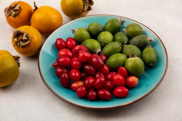 Vista superior de feijoas frescas com cerejas da cornalina em um prato azul com caquis isolados em um fundo cinza