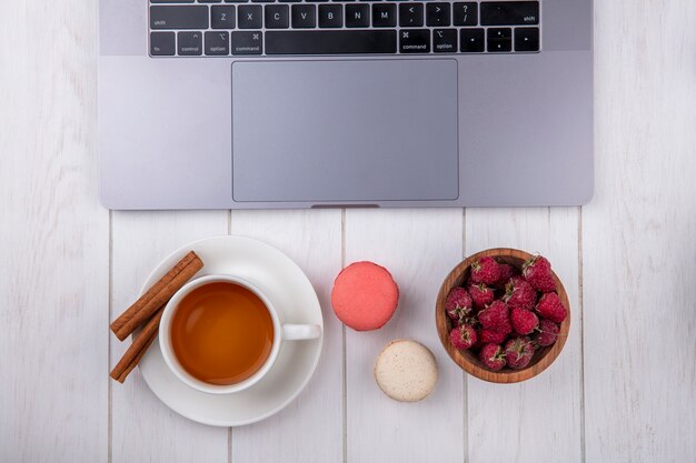 Vista superior de framboesas com uma xícara de biscoitos de chá e um laptop em uma superfície branca