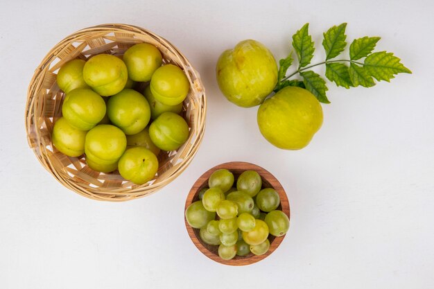 Vista superior de frutas como ameixas na cesta e pluots verdes com tigela de uvas brancas sobre fundo branco