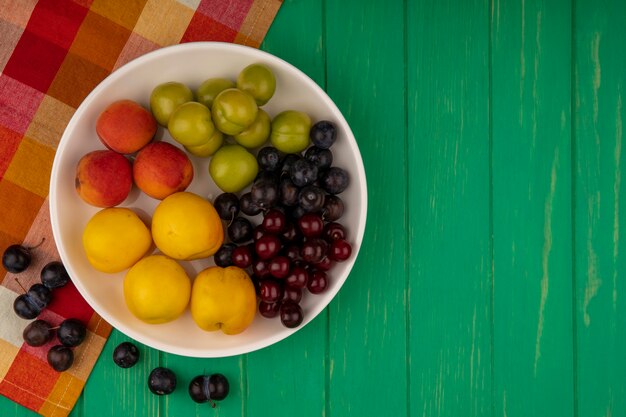 Vista superior de frutas como damascos, ameixas e cerejas em uma tigela com bagas de abrunheiro em tecido xadrez e sobre fundo verde com espaço de cópia