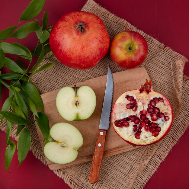 Vista superior de frutas como metade cortada maçã e romã metade com faca na tábua e maçã inteira e romã com folhas de saco na superfície vermelha