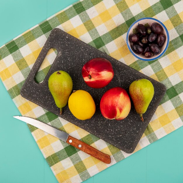 Vista superior de frutas como pêra, pêssego, limão na tábua de cortar com uvas e uma faca em pano xadrez sobre fundo azul