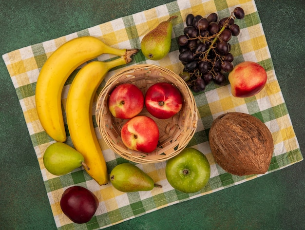 Vista superior de frutas como pêssego na cesta e uva pêra banana coco em pano xadrez sobre fundo verde