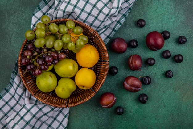 Vista superior de frutas como plumas de uvas e nectacots em uma cesta em tecido xadrez e padrão de pluots e bagas de uva em fundo verde