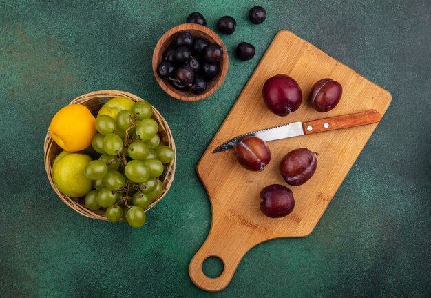 Vista superior de frutas como pluots com faca na tábua e pluots nectacots uva na cesta com bagas de uva na tigela e sobre fundo verde