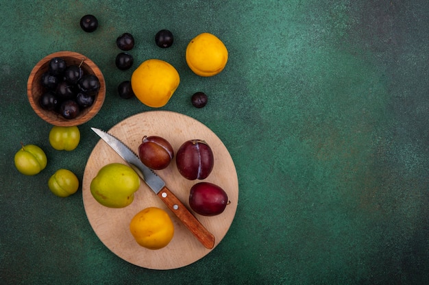 Vista superior de frutas como pluots e nectacota com faca na placa de corte e bagas de uva em uma tigela com nectacotes de ameixa e bagas de uva em fundo verde com espaço de cópia