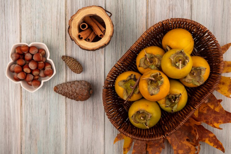 Foto grátis vista superior de frutas de caqui em um balde com avelãs em uma tigela com paus de canela em uma jarra de madeira em uma mesa de madeira cinza