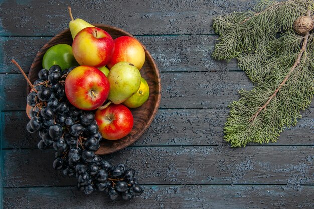 Vista superior de frutas distantes em uma tigela de uvas, peras, maçãs, limões, próximo a ramos de abeto com cone na mesa escura