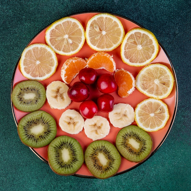 Vista superior de frutas fatiadas kiwi limão banana tangerina e cerejas vermelhas no prato no escuro