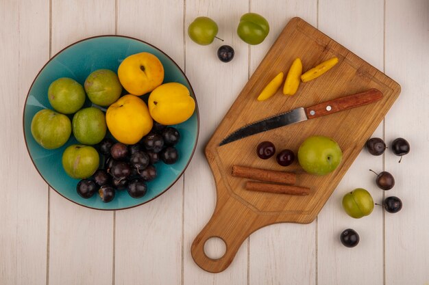 Vista superior de frutas frescas, como cereja verde, pêssegos e abrunhos frescos em uma tigela azul com fatias de pêssego em uma placa de cozinha de madeira com paus de canela com faca em um fundo branco de madeira