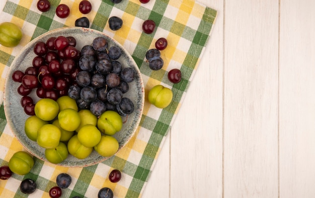 Vista superior de frutas frescas, como cherriessloes e ameixas de cereja verdes em uma tigela sobre uma toalha de mesa quadriculada em um fundo branco com espaço de cópia