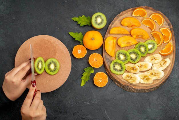 Vista superior de frutas frescas em uma bandeja de madeira e kiwis cortando à mão em uma tábua na superfície preta