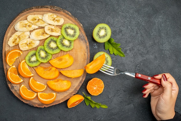 Vista superior de frutas frescas em uma bandeja de madeira segurando um garfo com uma fatia de kiwi no fundo escuro