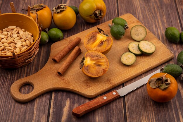 Vista superior de frutas meio caqui com fatias de feijoas com paus de canela em uma placa de cozinha de madeira com amendoim em um balde em um fundo de madeira