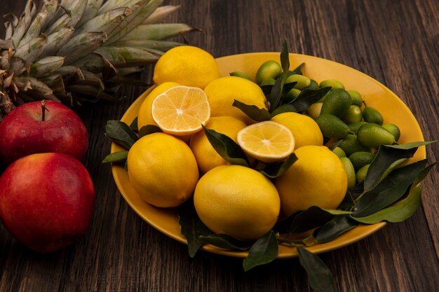 Vista superior de frutas ricas em vitaminas, como limões e kinkans, em um prato amarelo com abacaxi e maçãs vermelhas isoladas em uma parede de madeira