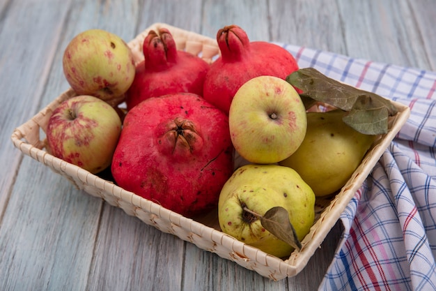 Vista superior de frutas saudáveis, como romãs, maçãs e marmelos em um balde em um pano xadrez em um fundo cinza