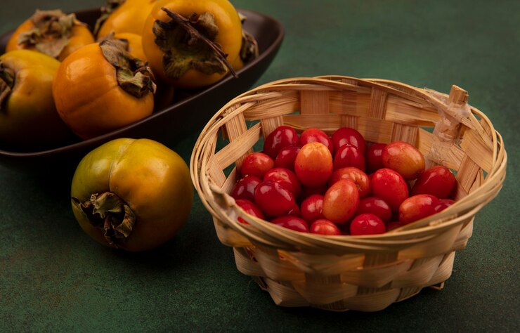 Foto grátis vista superior de frutos de caqui cor de laranja em uma tigela com cerejas da cornalina em um balde em uma superfície verde
