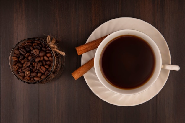 Vista superior de grãos de café em uma jarra de vidro com uma xícara de café com paus de canela em uma parede de madeira