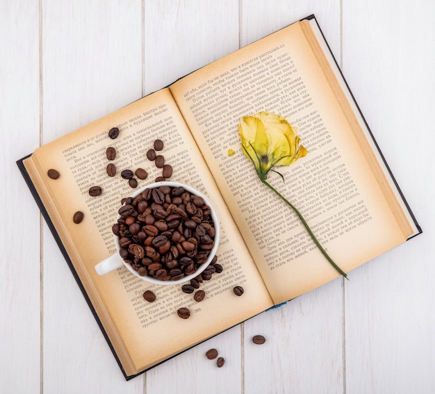 Foto grátis vista superior de grãos de café torrados escuros em um copo branco com uma flor em um fundo branco de madeira