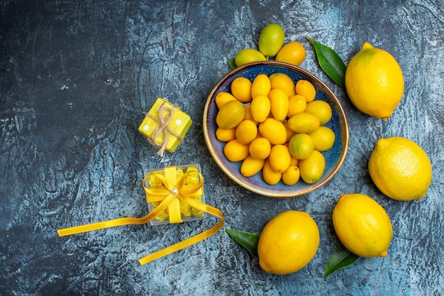 Vista superior de kumquats orgânicos em um prato azul, limões frescos com folhas e caixas de presente amarelas em fundo escuro