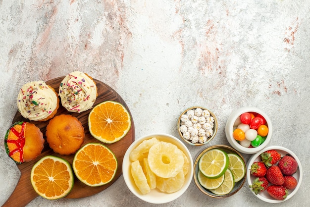 Vista superior de longe frutas cítricas no tabuleiro tigelas de morangos, limão, doces e abacaxis secos e fatias de laranja e biscoitos na tábua de madeira