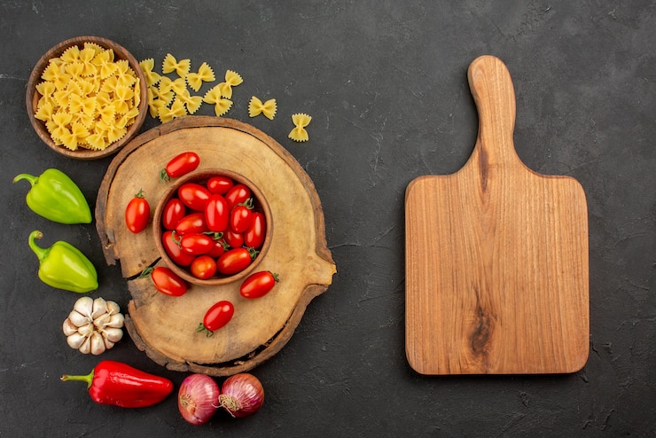 Foto grátis vista superior de longe macarrão e tomate tábua de cortar tigela marrom de tomates na tábua de madeira macarrão e cebola pimentão e alho na mesa