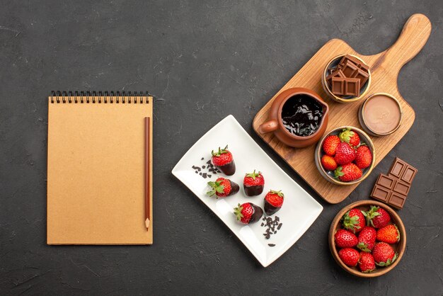 Vista superior de longe, morangos com chocolate, morangos com cobertura de chocolate, chocolate e tabuleiro de cozinha com creme de chocolate e morangos ao lado do caderno e do lápis