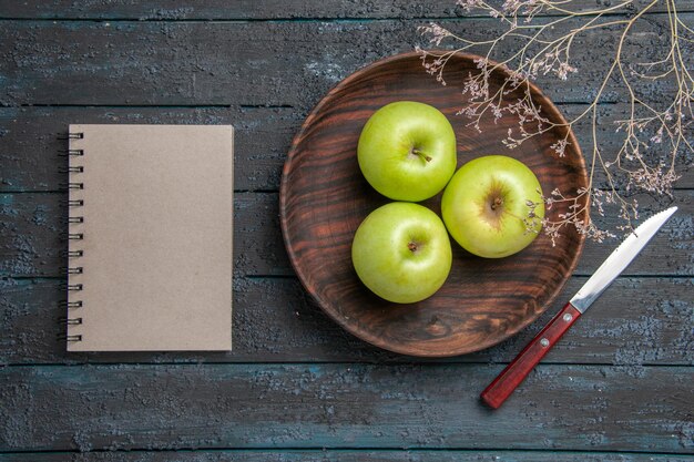 Vista superior de longe. Placa de maçãs Placa de madeira com maçãs apetitosas ao lado de um caderno cinza e galhos de árvores na superfície escura