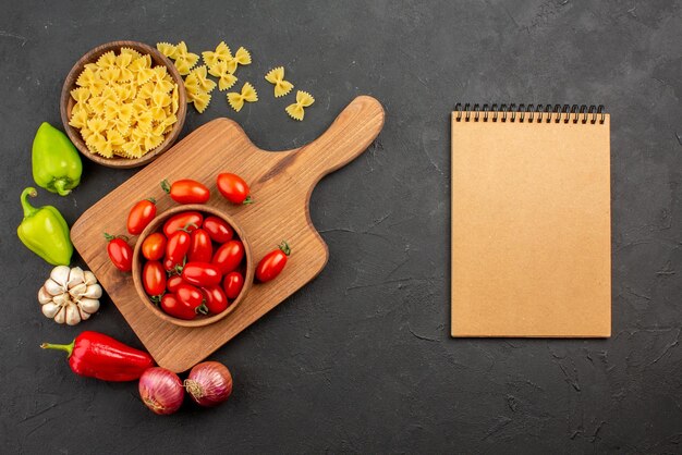 Vista superior de longe tomates e especiarias pimentão cebola alho e tigela de macarrão ao lado da tigela de tomates na placa de madeira e caderno de creme na mesa